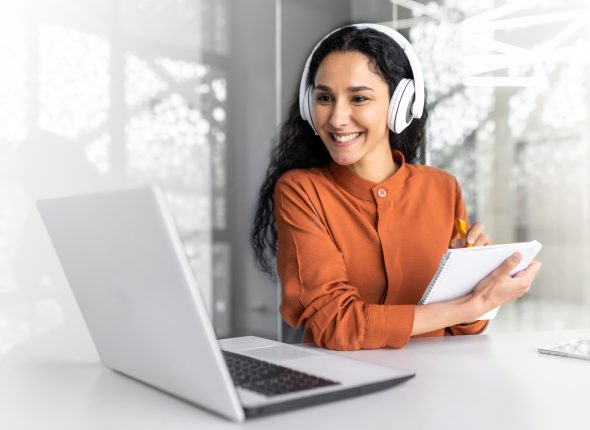 Latin american business woman with curly hair and headphones watching online training course at workplace, woman writing information happy and satisfied with the results of professional development